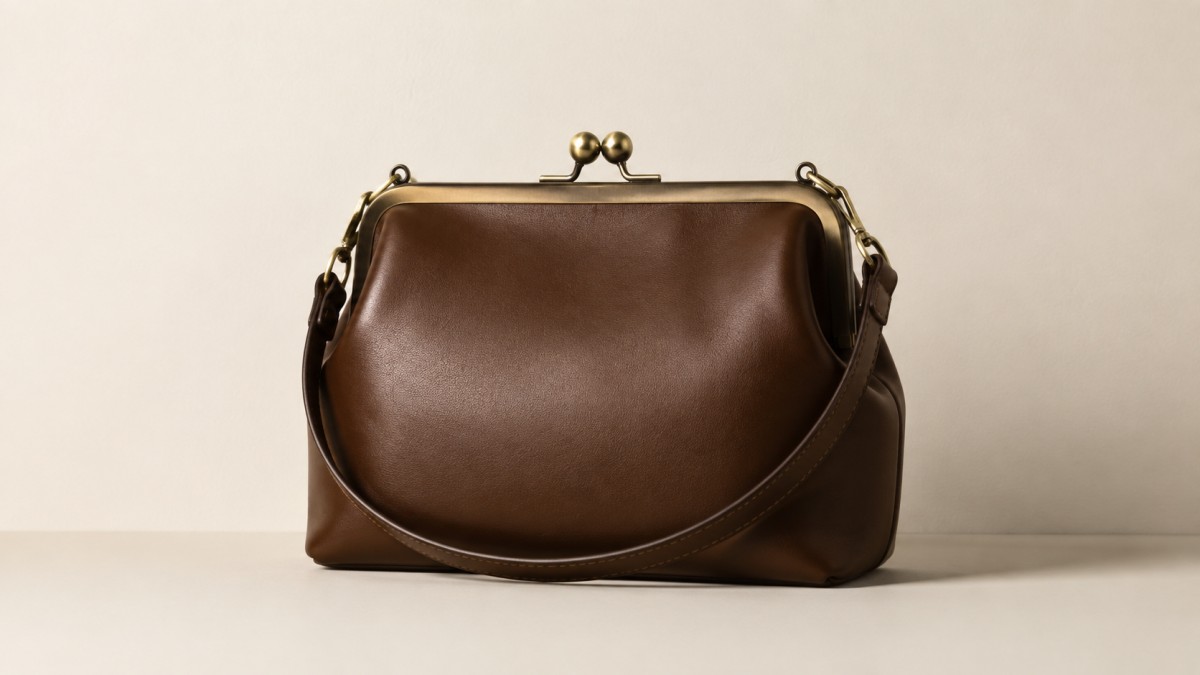 Leather frame bag with a kiss-lock closure against a plain warm off-white wall